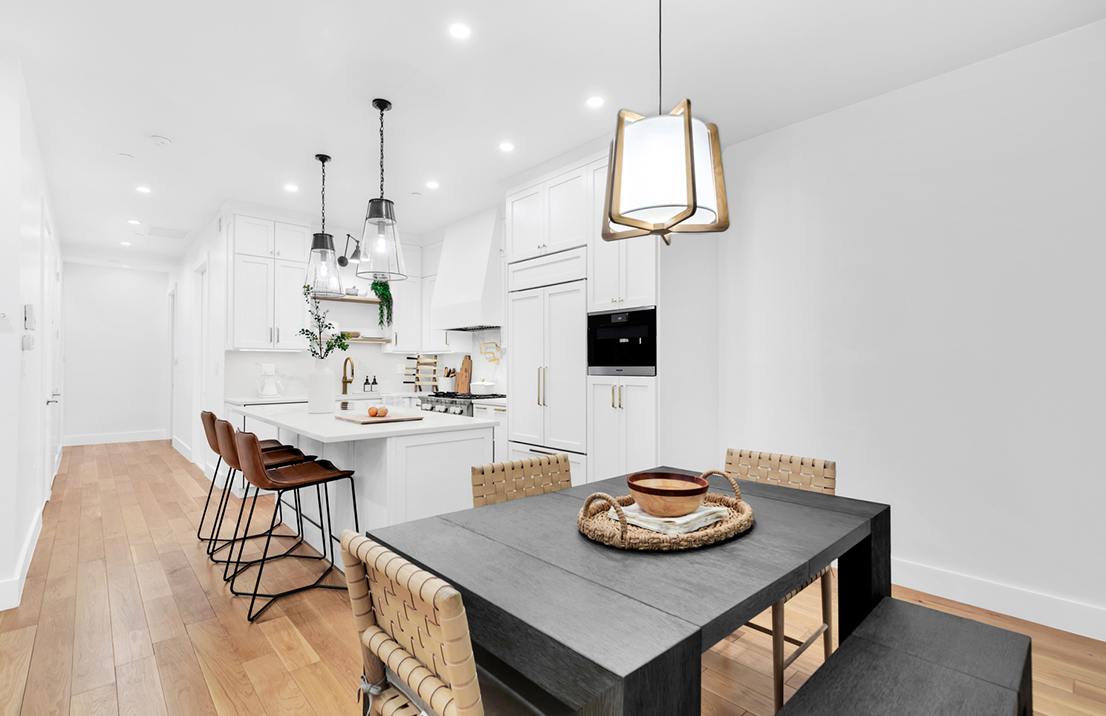 Remodeled kitchen all white, with recessed lighting and dangling light pendants, and an island with brown chairs.